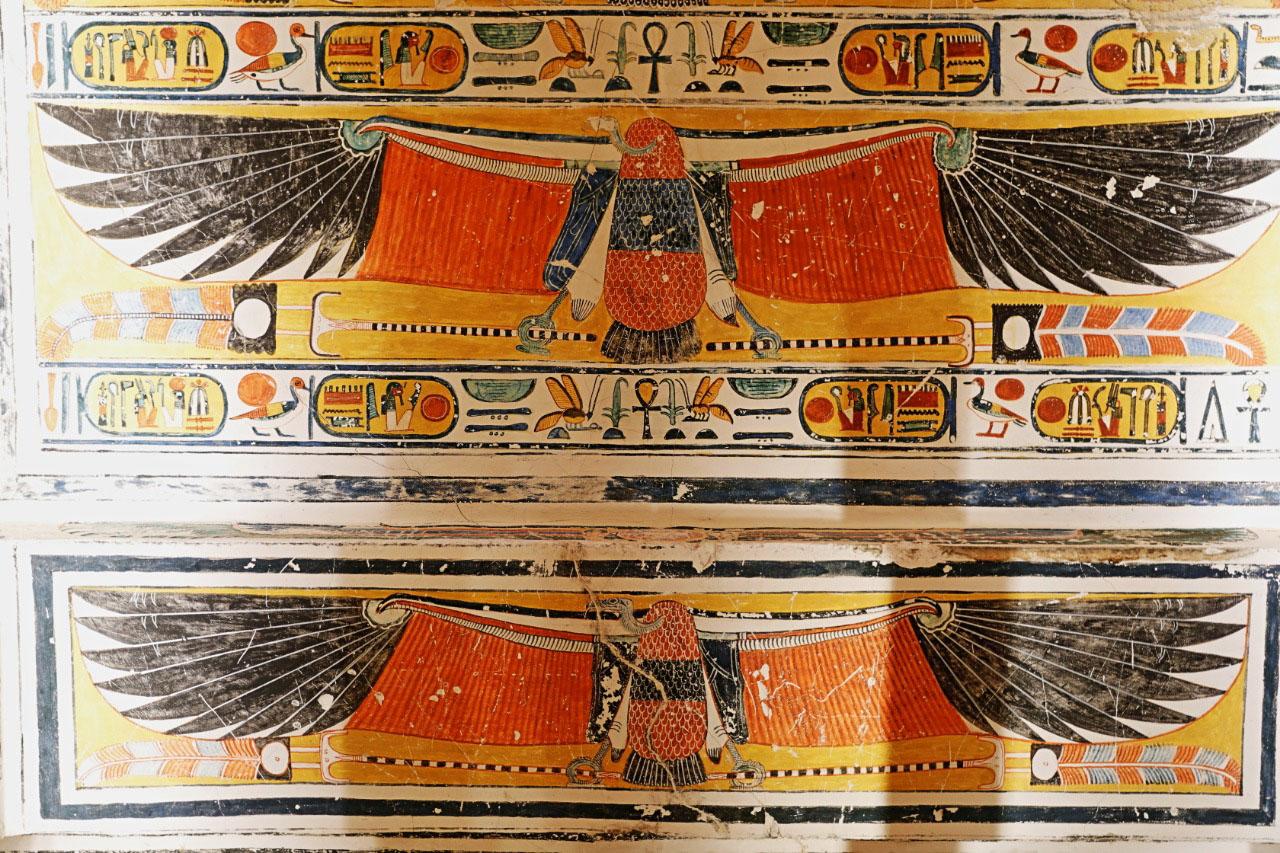 Detail of a painted winged scarab frieze on a tomb wall, flanked by cartouches and deities.