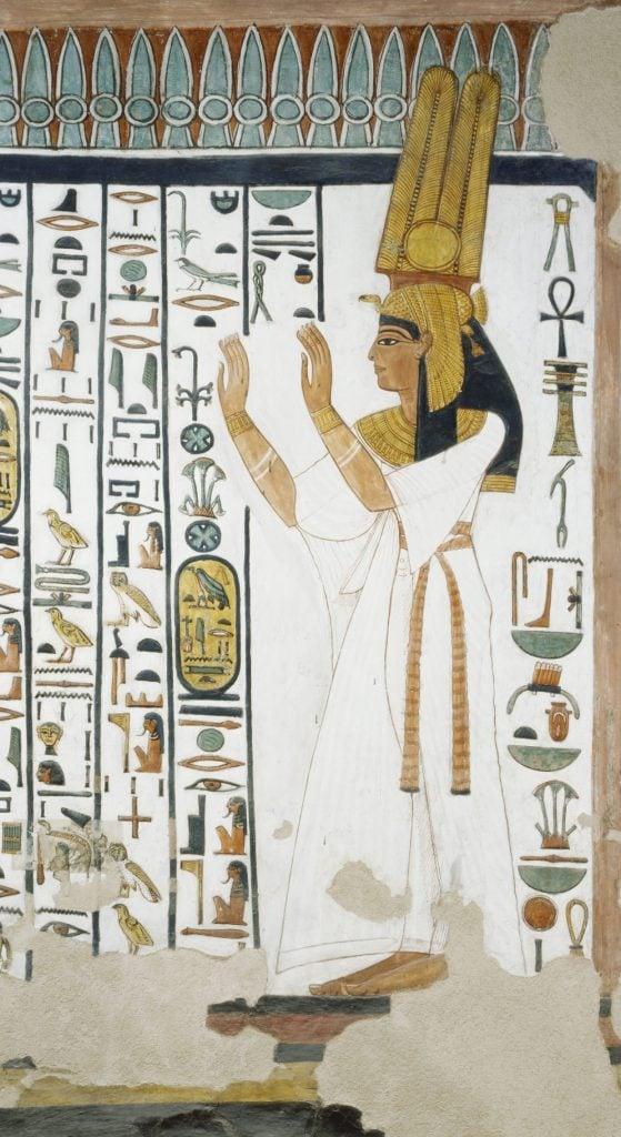 Nefertari in white gown and plumed crown raising hands before a column of hieroglyphs.