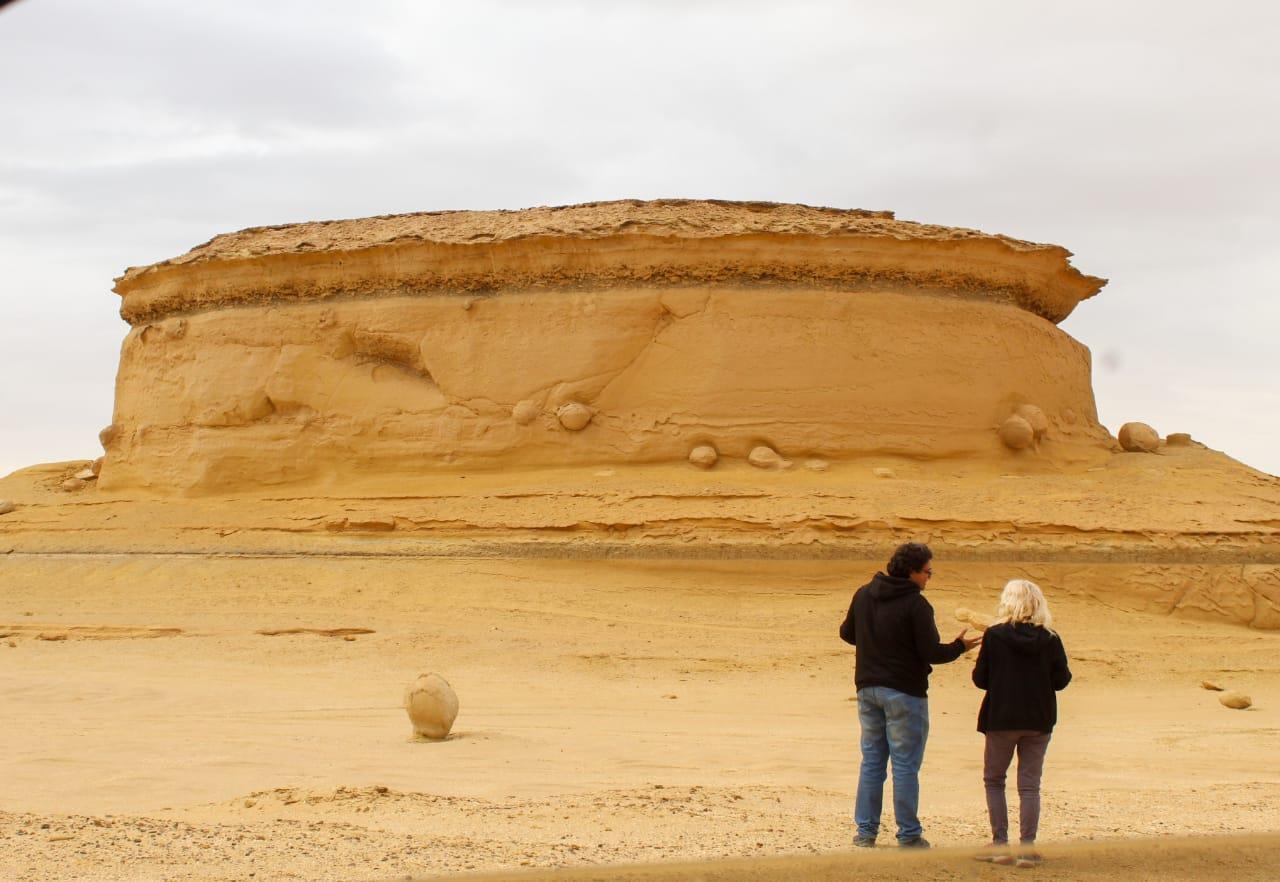 Two visitors examining a fossil beside a layered sandstone outcrop at Wadi Al-Hitan in the Fayoum desert