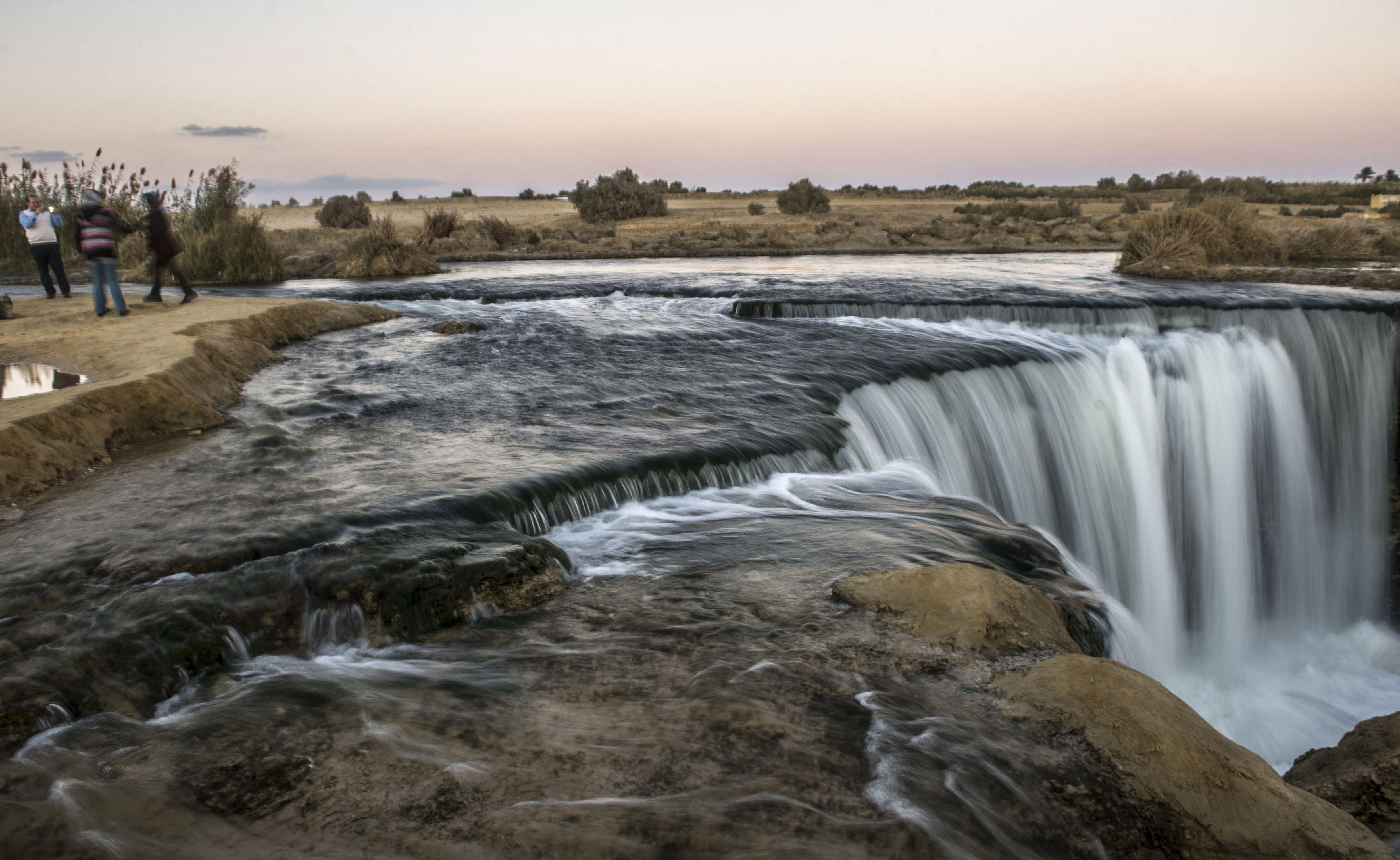 Visitors watching a wide cascade tumble over a desert cliff at dusk
